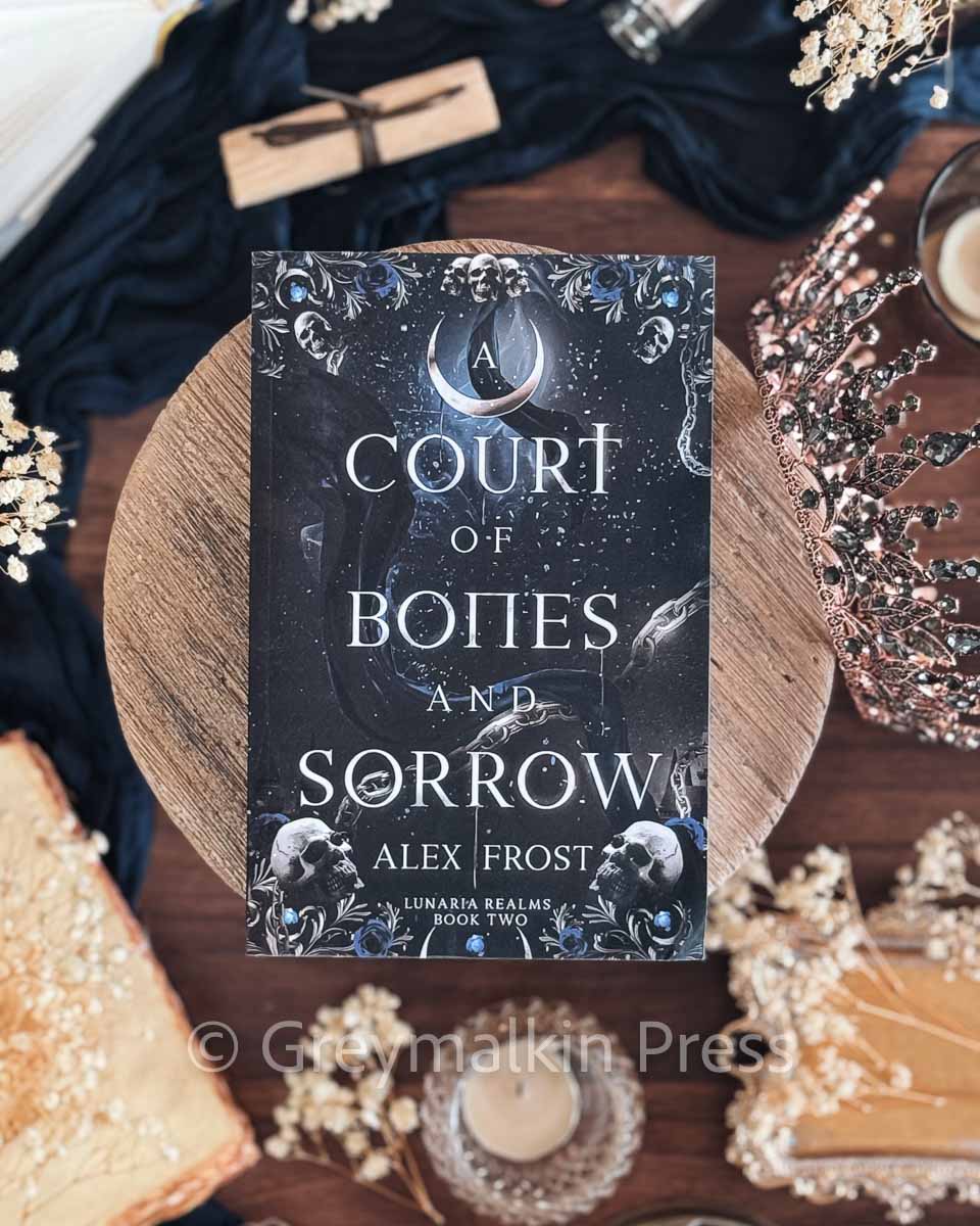 Book titled 'A Court of Bones and Sorrow' by Alex Frost on a decorative surface with flowers and candles.