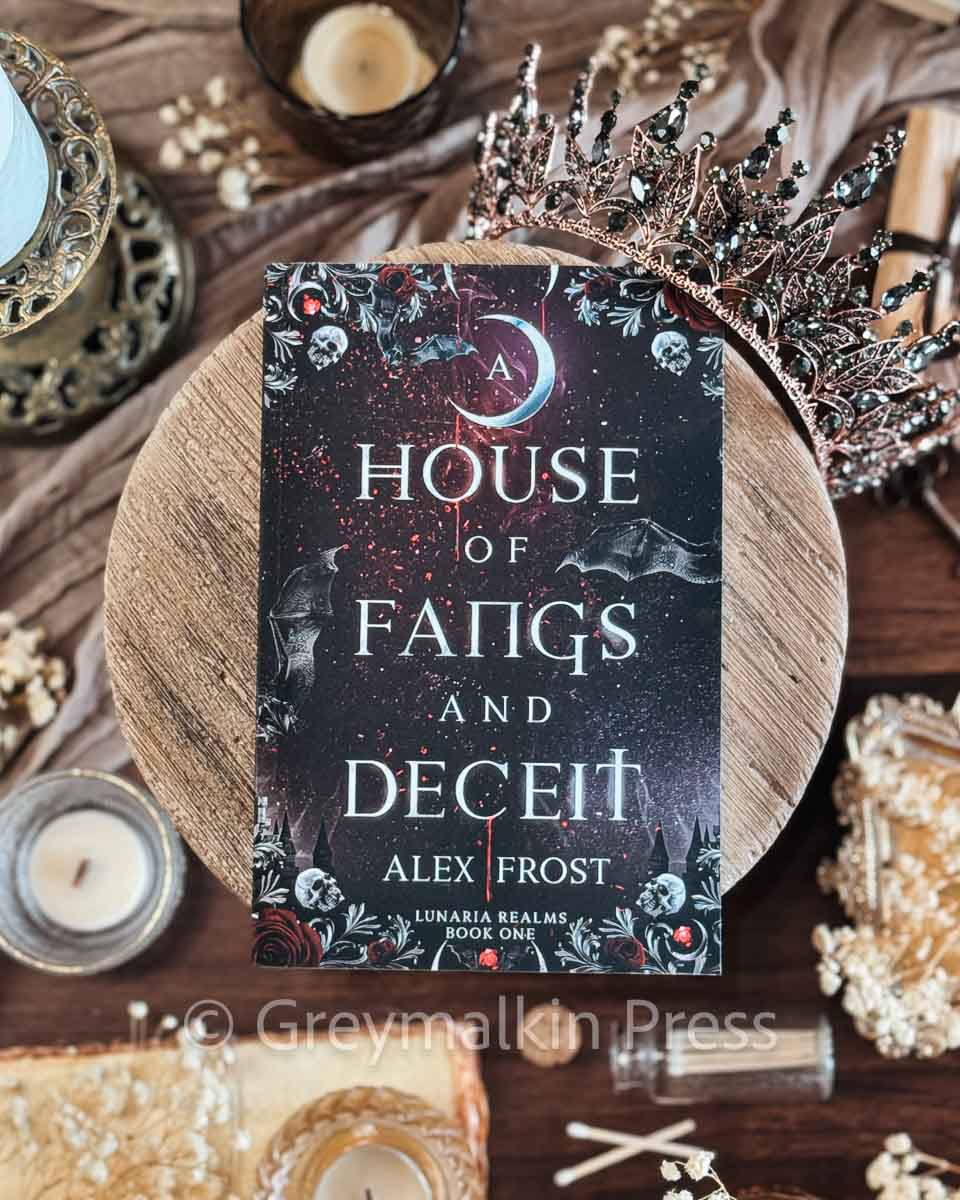 Book titled 'The House of Fangs and Deceit' by Alex Frost on a wooden surface with decorative items.