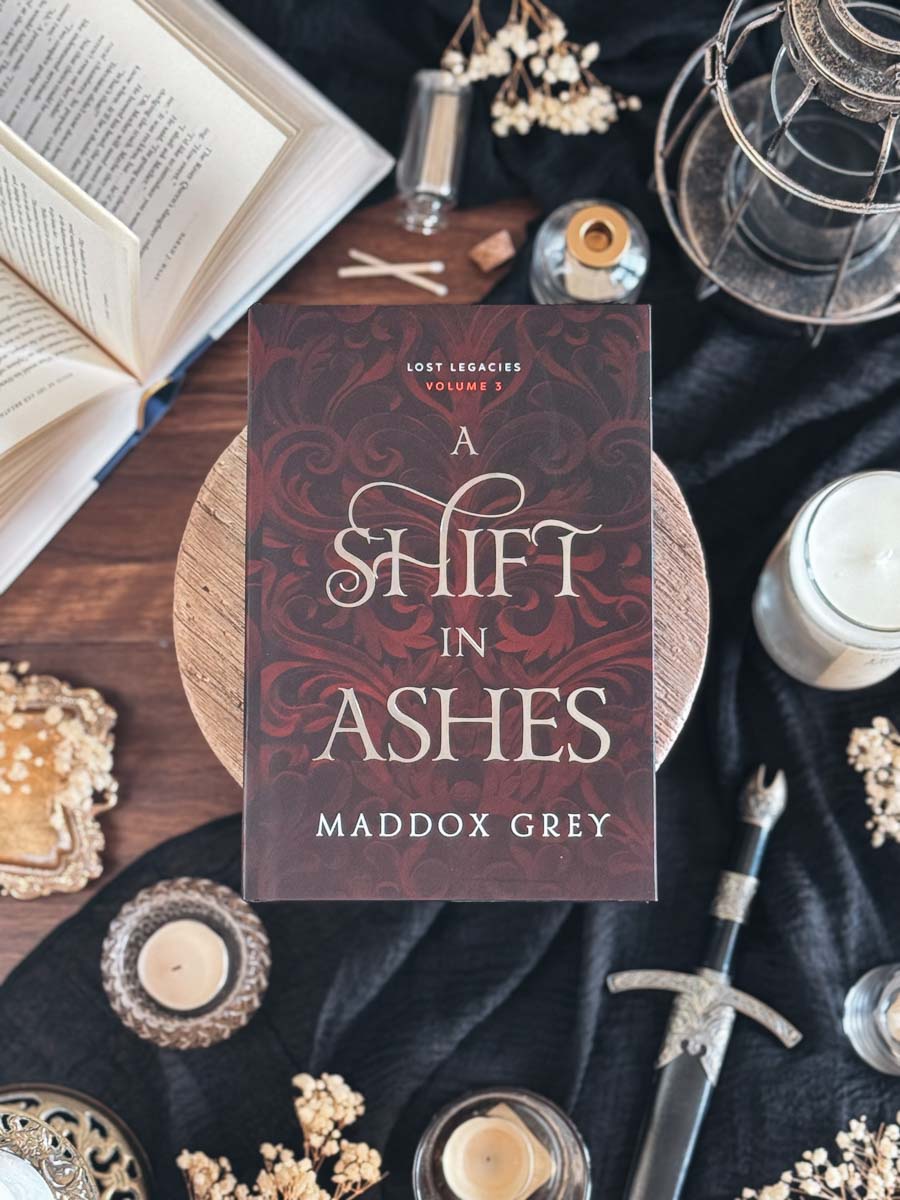 Book cover of 'A Shift in Ashes' by Maddox Grey on a dark fabric background with decorative items.