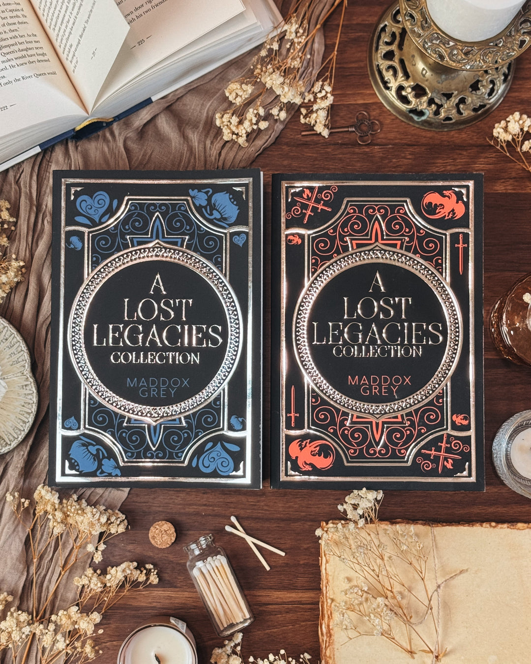 Two 'A Lost Legacies Collection' books by Maddox on a wooden surface with candles and dried flowers.